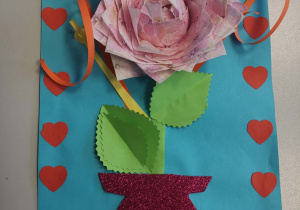 To ręcznie wykonana dekoracja przedstawiająca trójwymiarowy kwiat z papieru umieszczony na niebieskim tle ozdobionym czerwonymi serduszkami. Centralny kwiat ma warstwowe, różowe płatki ułożone tak, by wyglądały jak prawdziwe, a obok niego znajdują się dwa zielone liście o postrzępionych brzegach. Od łodygi odchodzą zwinięte w spirale wstążki w kolorze pomarańczowym i żółtym. Całość „stoi” w czerwonym, brokatowym wazonie wyciętym z błyszczącego papieru.