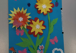 Jest to laurka na Dzień Kobiet. Praca plastyczna przedstawia trzy kolorowe, warstwowe kwiaty na zielonych łodygach, umieszczone na niebieskim tle. Wokół znajdują się różowe i niebieskie motylki, brokatowe serduszka, białe kwiatki oraz błyszczące kryształki. Na dole jest zielona „trawa” z papieru i dwie naklejki kotów. Całość jest bardzo kolorowa i wesoła.