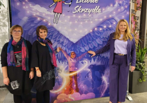 Na zdjęciu stoją trzy kobiety przed barwną, bajkową scenografią. Tło przedstawia dziewczynkę z anielskimi skrzydłami, otoczoną światełkami i pastelowymi chmurami, a poniżej wkomponowane jest zdjęcie dziecka z rozłożonymi ramionami, jakby samo miało skrzydła. Obok widnieje nazwa „Liliowe Skrzydła – Fundacja im. Lilianki Błasiak”. Całość tworzy ciepłą, uroczystą atmosferę wydarzenia związanego z fundacją i pamięcią o Liliance. Te kobiety - to mama Lilianki oraz autorki wspomagające Marta Januszewicz i Agnieszka Adamczyk.