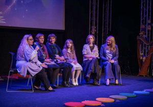 Na zdjęciu widać panel sześciu osób siedzących na scenie podczas rozmowy lub prezentacji. Jedna z nich trzyma mikrofon i prowadzi dyskusję. Obok stoi harfa, a tło tworzy duży ekran z abstrakcyjną grafiką. Przed sceną leżą kolorowe, okrągłe maty, które dodają wydarzeniu lekkiego, przyjaznego charakteru.