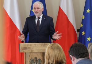 Na zdjęciu widać mężczyznę przemawiającego z mównicy ozdobionej polskim godłem. Za nim stoją flagi Polski i Unii Europejskiej, co nadaje scenie oficjalny, państwowy charakter. Przed mównicą siedzi publiczność, a całość wygląda jak formalne wystąpienie lub konferencja w instytucji rządowej. Przedstawionym mężczyzną jest Pan Janusz Brzozowski - nasz Łódzki Kurator Oświaty.