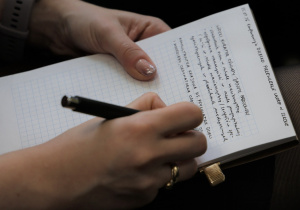 Na zdjęciu widać osobę robiącą odręczne notatki w zeszycie w kratkę. Pisze czarnym długopisem, a na dłoni ma pierścionek i smartwatch. Notatki dotyczą konferencji o dziecku przewlekle chorym w szkole — zapisane są daty, nazwiska i punkty z wystąpienia. Ujęcie jest bliskie, skupione na dłoni i zeszycie, co podkreśla roboczy, merytoryczny charakter sytuacji.