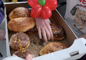Zdjęcie przedstawia dziecięcą rękę ozdobioną balonowym kwiatkiem. Dłoń sięga po słodką bułkę.