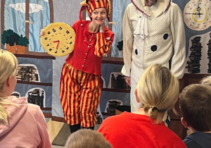 Zdjęcie przedstawia aktorów teatralnych kobietę i mężczyznę. Kobieta trzyma zabawkowy zegar. Widać też zgromadzone na widowni dzieci.