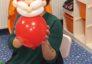 Pani siedzi i pokazuje balonik zwinięty w kształcie czerwonego Mikołaja