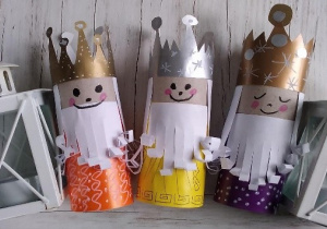 Trzy Mikołaje wykonane z papieru z koronami na głowie, obok stojące obok świecznikami w kształcie latarek