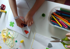 Zdjęcie przedstawia ręce dziecka, które ozdabia wykonaną z papieru torebkę. Nakleja kolorowe elementy kwiatków, koniczynkę, serduszka. Obok torebki znajdują się kolorowe paski pocięte z papieru. Na stole stoi pudełko z różnokolorowymi kredkami.