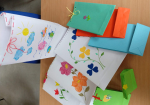 Torby papierowe ozdobione kolorowymi, narysowanymi przez dzieci kwiatami. Na torbie ustawionej pionowo, widać morskie motywy: piankowe rybki, ośmiornica, dorysowane chmurki i słońce. W dziurki znajdujące się na górnym brzegu torebki, wpleciony jest czerwony sznurek, zawiązany na kokardkę. Na dole zdjęcia stoją małe, zielone torebki, ozdobione serduszkami.