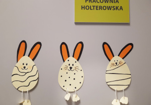 Trzy beżowe pisanki w czarne wzorki z króliczymi uszami i nóżkami na sznurku.