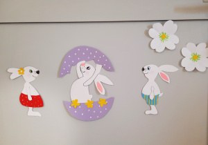 Trzy białe króliki. Jeden wszedł do pisanki i podnosi jej górną część, dwa pozostałe przyglądają się koledze siłaczowi.