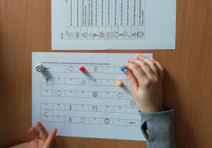 Na zdjęciu widoczna jest plansza edukacyjnej gry, pionki, kostka i dłonie uczniów. Obok znajduje się lista z pytaniami.