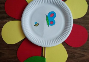 kwiat wykonany z talerzyka papierowego z żółtymi i czerwonymi płatkami. Posrodku narysowany motyl i pszczółka.
