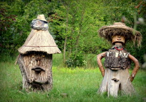 Zdjęcie zrobione w arboretum w Rogowie. Na zielonej trawie, na tle drzew stoją dwa ule. Ul stojący z lewej strony zrobiony jest z pnia drzewa, z dorobionymi oczami, nosem i ustami z gałązek oraz nasadzonym na górze w kształcie słomianej strzechy, kapeluszem. Z prawej strony znajduje się ul w kształcie człowieka, ze słomianym, podobnym do sombrero kapeluszem i czerwonymi koralami na szyi.