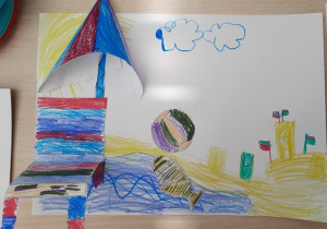 Rysunek dziecka "Plażing" - kolorowy leżak nad morzem, nad nim parasol plażowy, dużo piasku i zbudowany z niego zamek, w wodzie pilka i ryba,