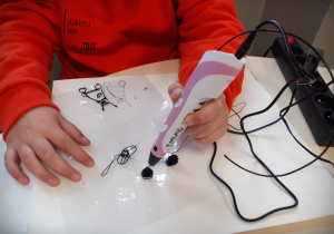 Dziecko w czerwonej bluzie wykonuje długopisem 3D, przestrzenną pracę przedstawiającą ufoludki.