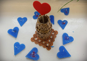 Na środku zdjęcia wazonik ozdobiony pestkami, w wazoniku czerwone serce na patyczku. Wazonik stoi na podkładce wykonanej z drewnianych kółeczek. Dookoła wazonika niebieskie świeczki w kształcie serc.