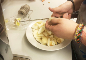 Na zdjęciu ręce dziewczyny, sznurek, obieraczka do warzyw i biały blat. Dziewczyna kroi nożem jabłka na małe kawałki.