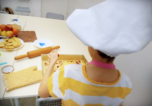 Na zdjęciu dziecko w kucharskiej czapce i koszulce w żółto- białe paski. Dziecko nakłada nadzienie na ciasteczka, obok leży rękawica kucharska, wałek, niebieska świeczka, owoce i herbatniki. W tle szare krzesła i drzwi.