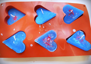 Na zdjęciu czerwona, silikonowa foremka z sześcioma otworami w kształcie serc. W każdym z otworów niebieska świeca.