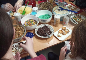 Jasny stol, na nim serek mascarpone, krem czekoladowy, orzechy w miseczce, owoce w słoiku i cukiernicze, kolorowe posypki. Dzieci siedzą przy stole i ozdabiają babeczki.