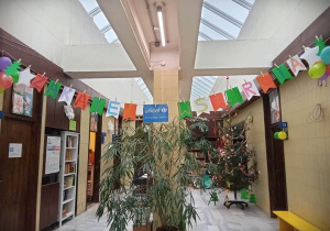 Na zdjęciu korytarz, na ziemi stoją zielone kwiaty, nad nimi wisi napis na kolorowych kartkach Zakątek wsparcia, oraz kartka z napisem Unicef dla każdego dziecka. Po lewej stronie stoi biały regal z książkami, po prawej choinka. W tle kolejny regal z książkami i wisząca na ścianie różowa skrzyneczka.
