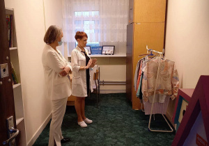 Na zdjęciu pokój pomalowany jasną farbą, po lewej stronie dwie kobiety w białych strojach. Po prawej stronie wieszak z ubraniami i brązowa szafa. W tle okno z firanką, na parapecie kolorowa praca w ramce i kartka z napisem UNICEF.