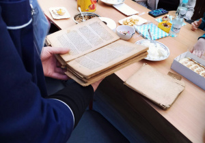 Na zdjęciu ręce kobiety trzymające starą książkę, w tle stol z produktami spożywczymi. Przy stole siedzą dzieci.