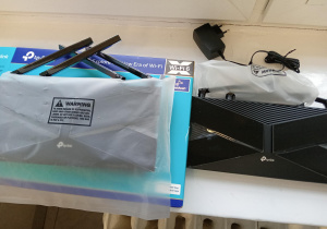 Na zdjęciu dwa czarne routery tp link, zasilacz i niebieskie pudełko.